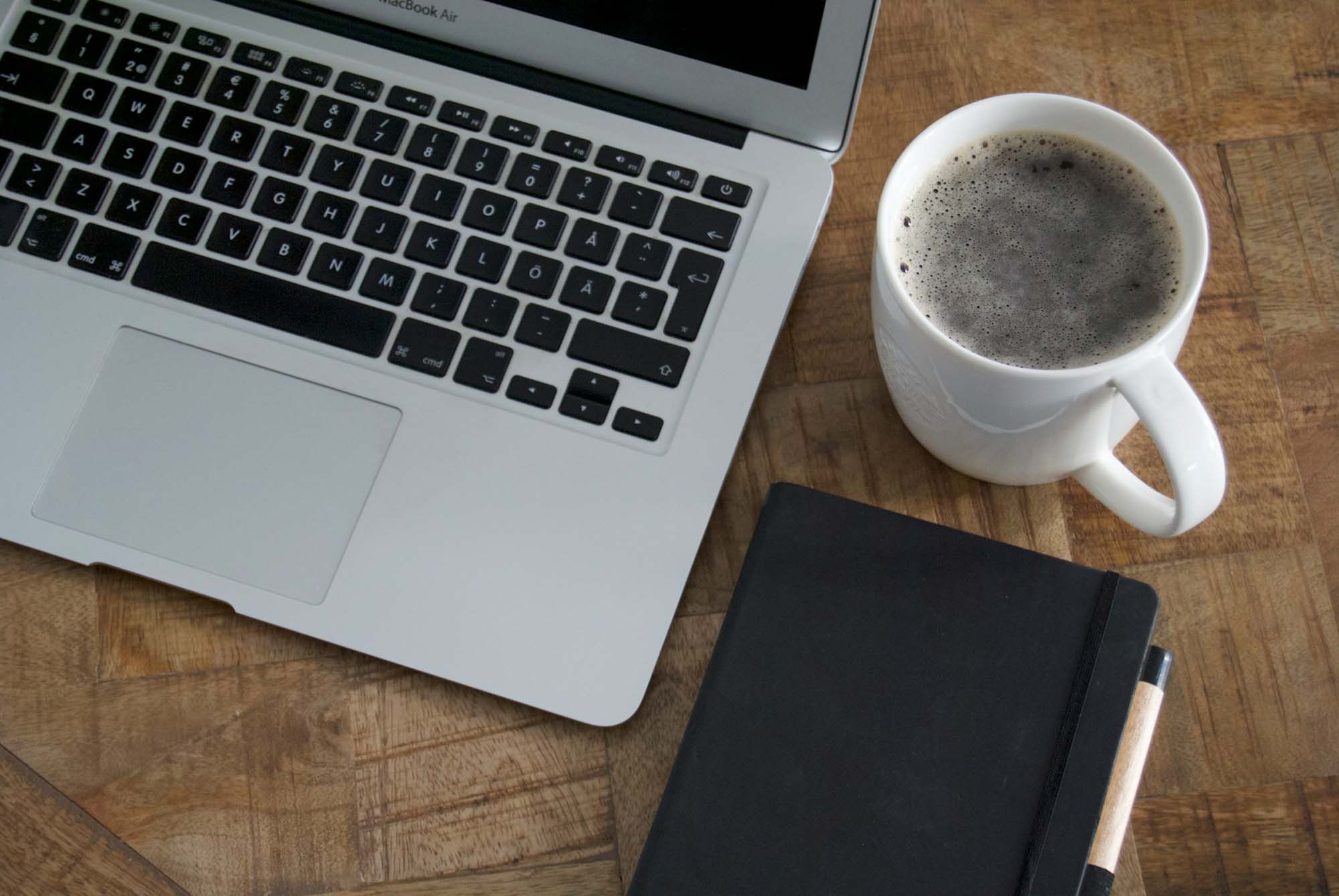 An open laptop, a cup of coffee, and a notebook sit on a table